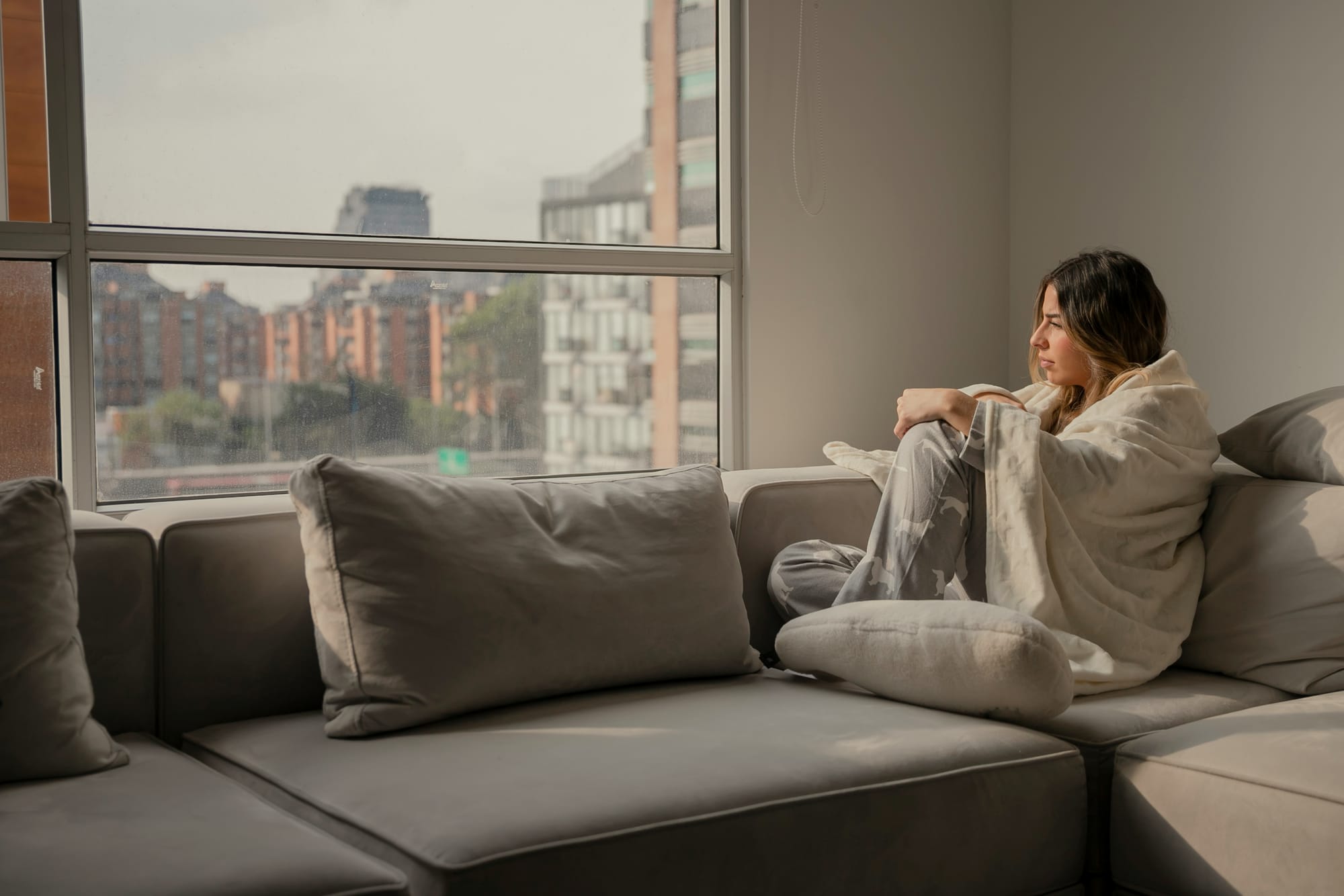 Woman sitting on a sofa wrapped in a blanket, gazing out the window — symbolising fatigue and introspection linked to high cortisol and impaired HPA axis function.