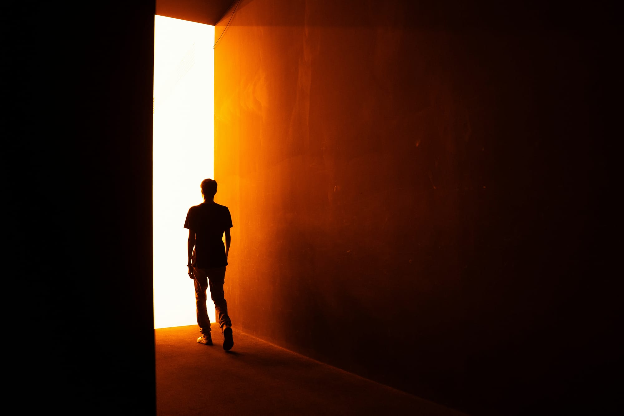 A silhouette of a person walking out of a dark, amber-lit hallway into a bright, glowing open doorway, symbolizing exiting the performance of caregiving and reclaiming reality.