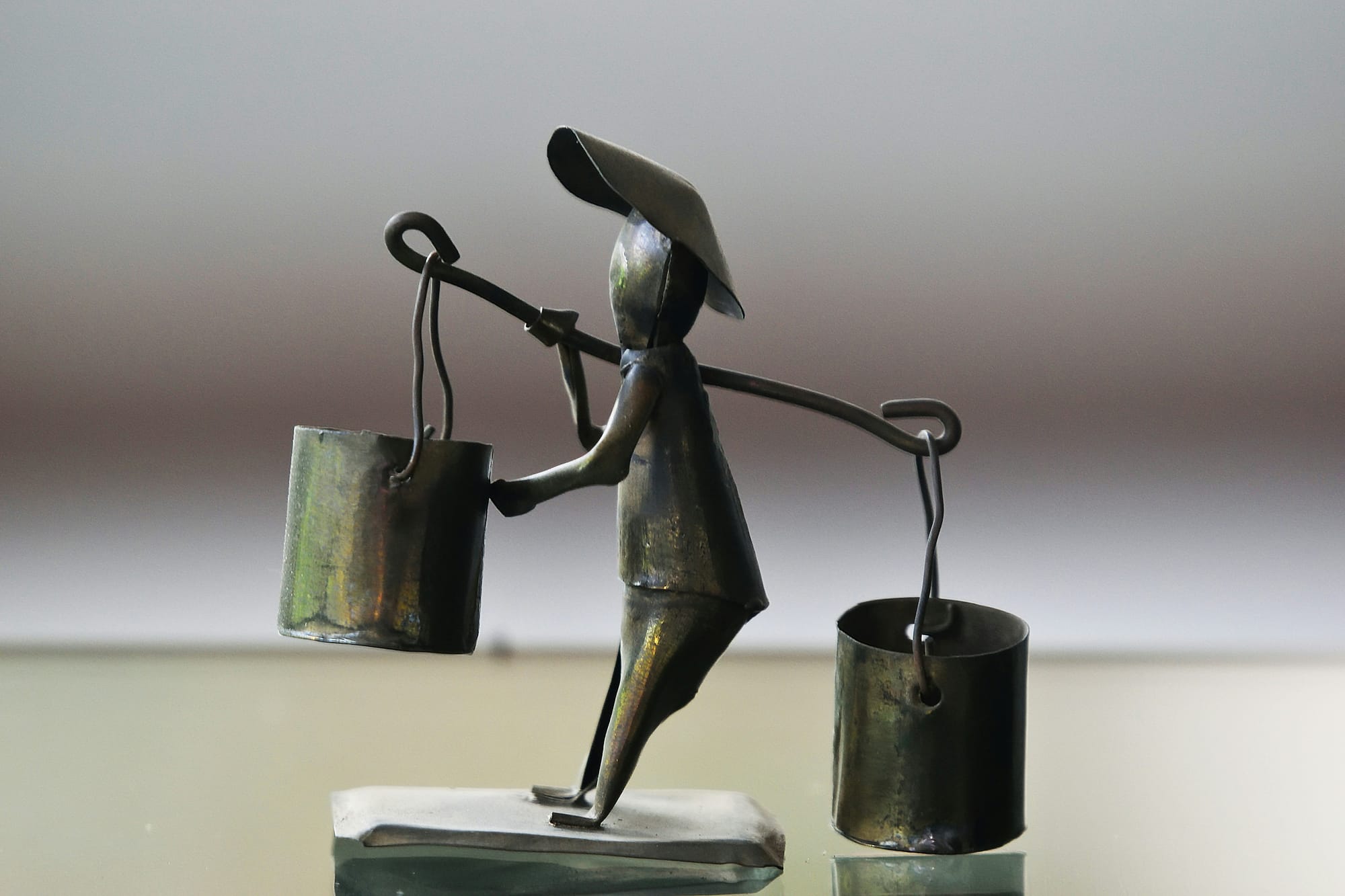 A weathered metal figurine carrying a shoulder yoke with two heavy buckets, representing the physical labor and heavy weight of caregiving.
