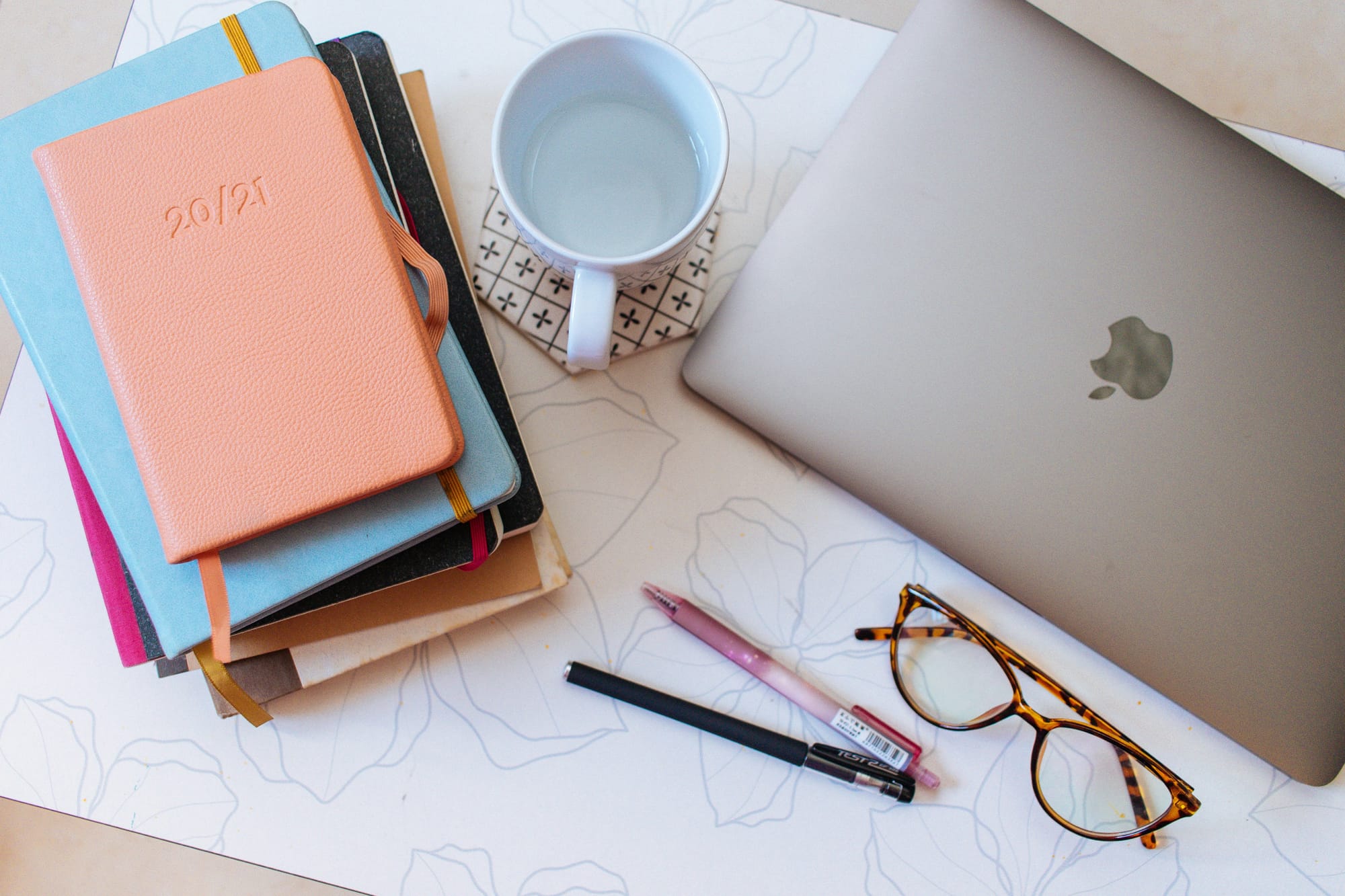 Stack of pastel notebooks, laptop, and glasses on a light floral desk — a calm workspace symbolizing structured, science‑rooted learning