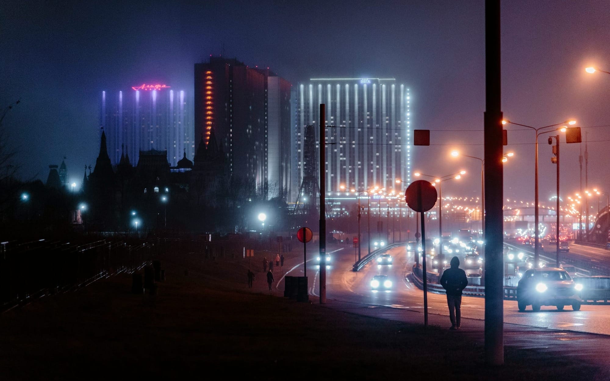 A moody nighttime view of a busy city highway with car headlights and tall, glowing neon skyscrapers, featuring a lone pedestrian silhouette walking along the dark roadside.
