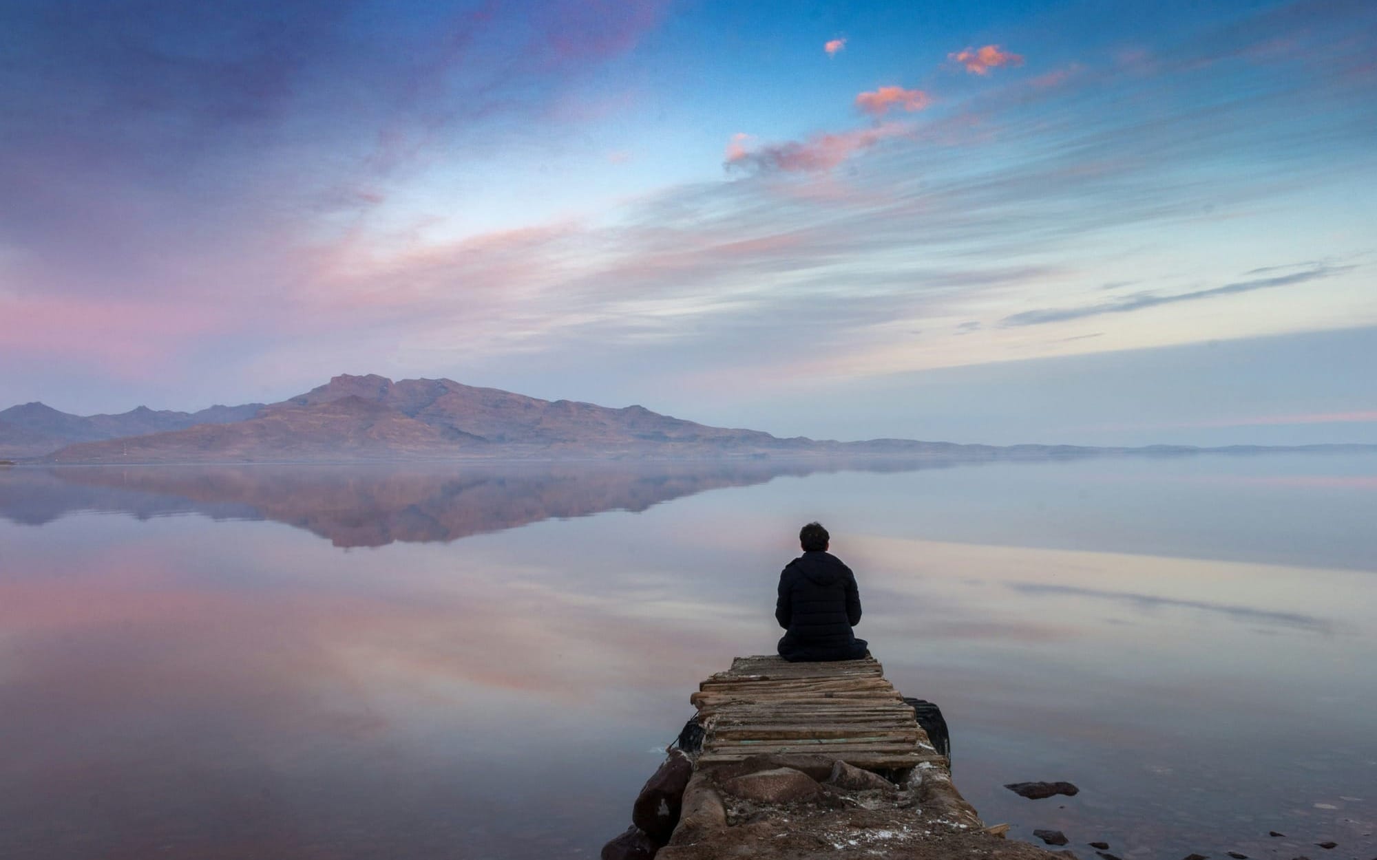 A silhouette of a lone person sitting quietly at the end of a wooden dock, facing a vast, calm lake reflecting a serene pink and purple sunset sky and distant mountains.