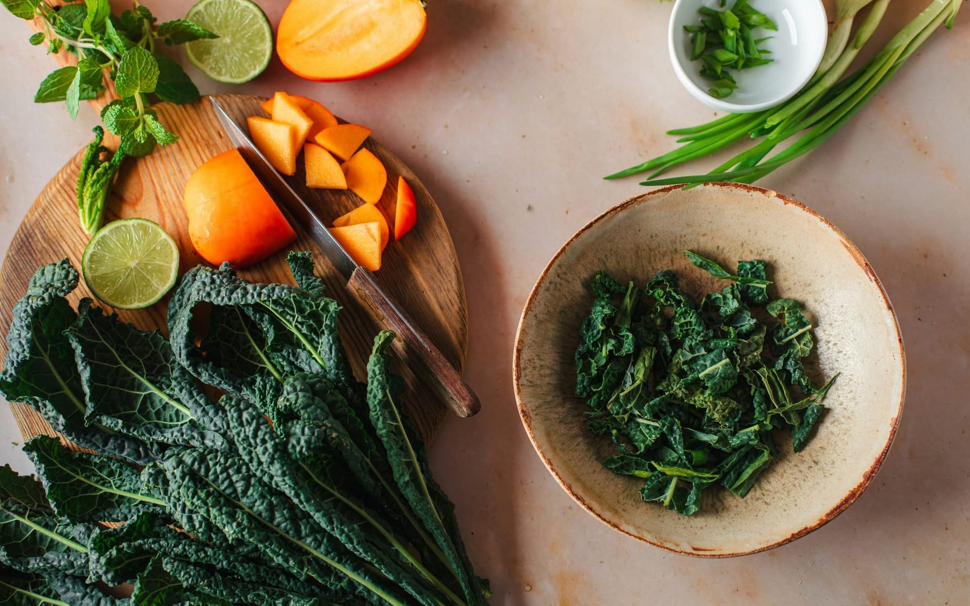 An overhead flat lay of dark green Lacinato kale, sliced orange persimmons on a wooden cutting board, and chopped greens in a ceramic bowl, representing magnesium-rich foods.