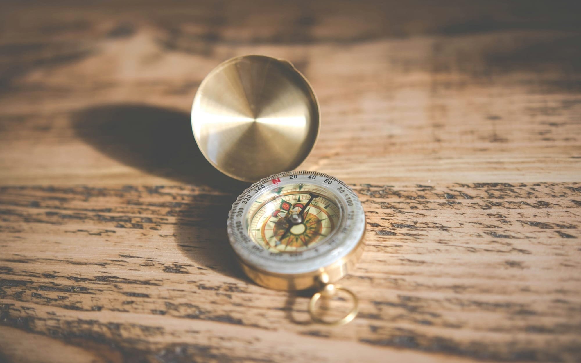 A close-up shot of an open vintage brass pocket compass resting on a warm, textured wooden table with soft focus.