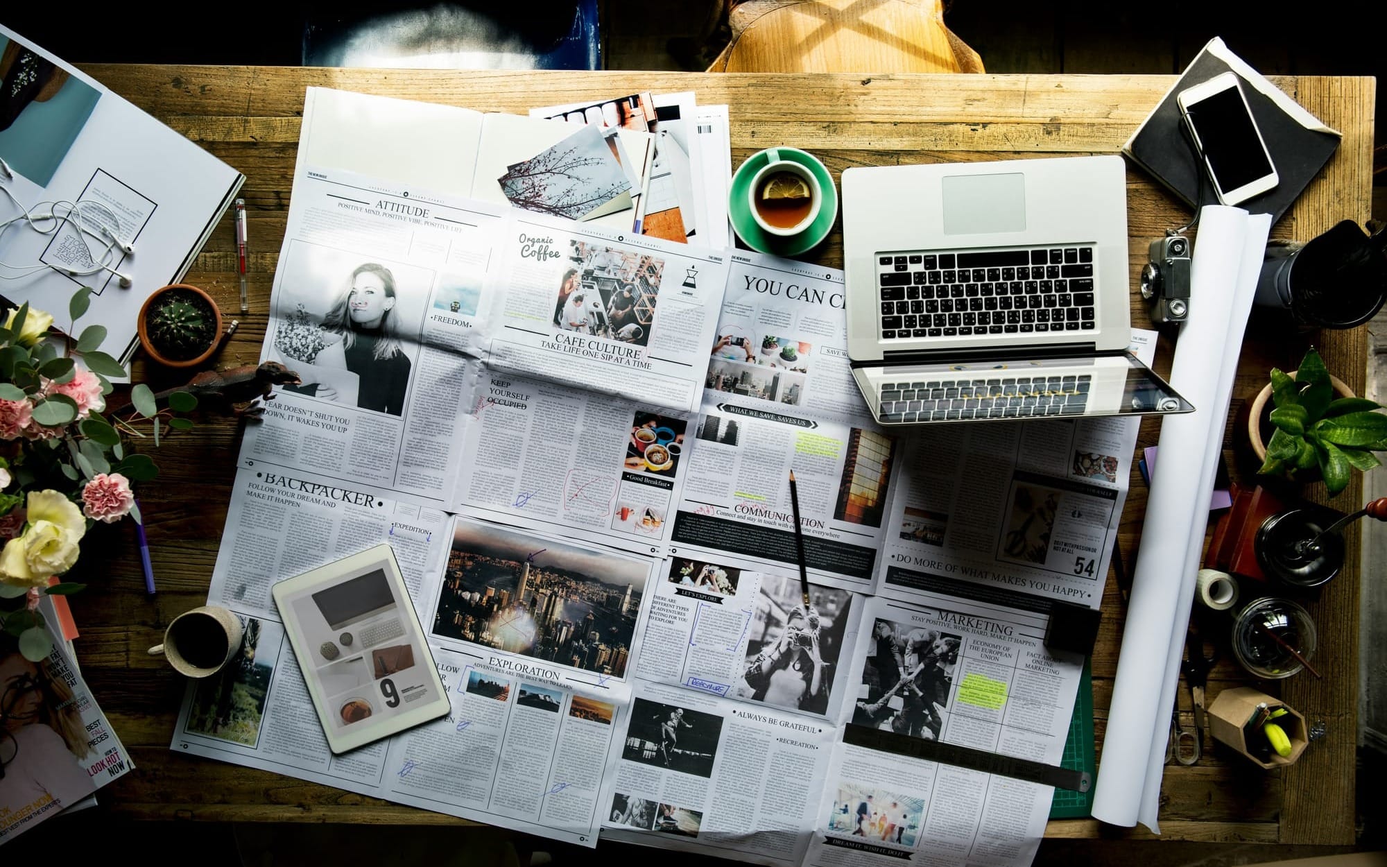 An overhead flat lay of a wooden desk covered in scattered newspapers, magazines, a laptop, and digital tablets, symbolizing the overwhelming media noise of modern diet culture.
