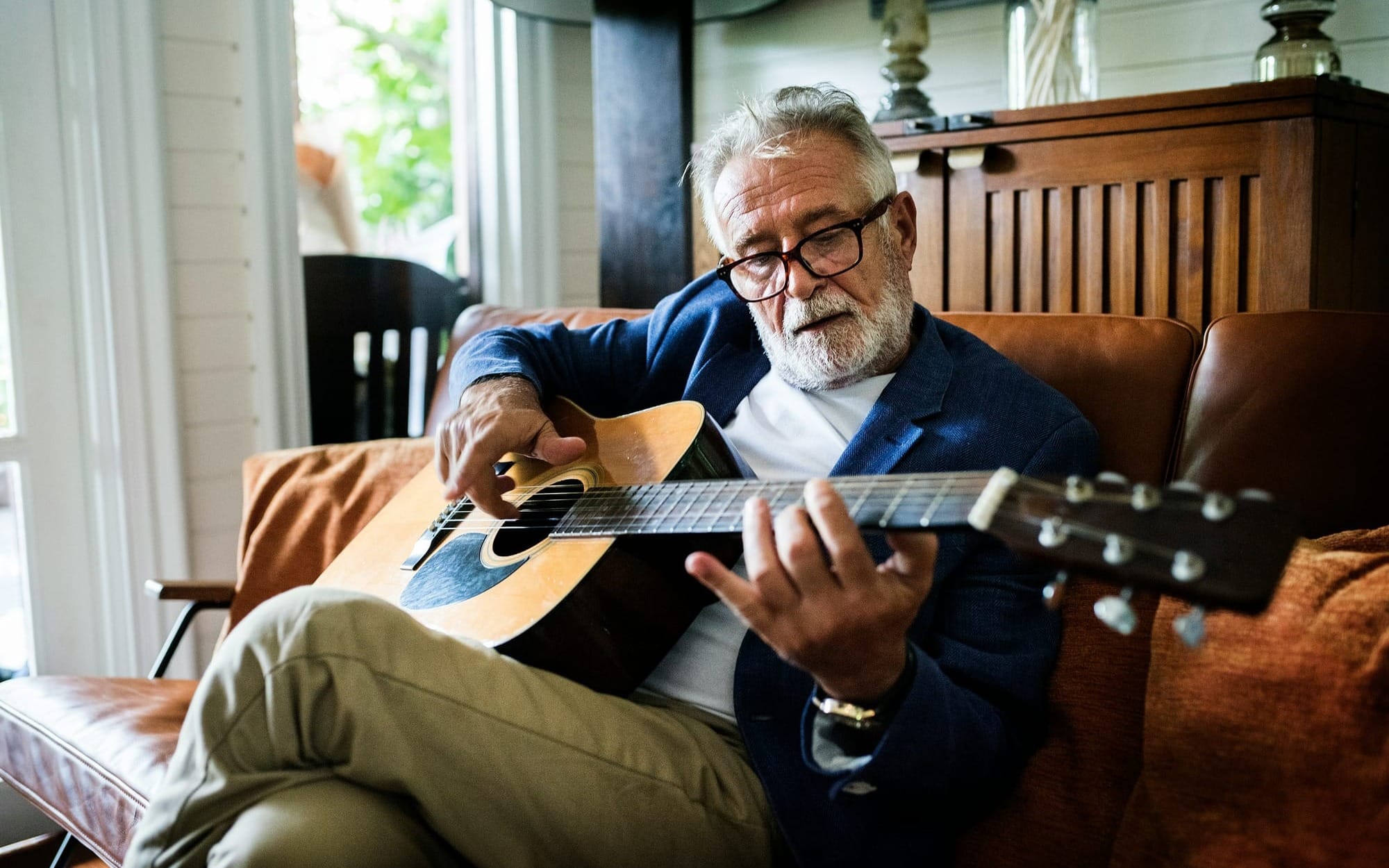 A senior man with glasses sitting on a leather sofa at home, deeply focused while playing an acoustic guitar, demonstrating baseline cognitive reserve and active neuroplasticity in healthy older adults.