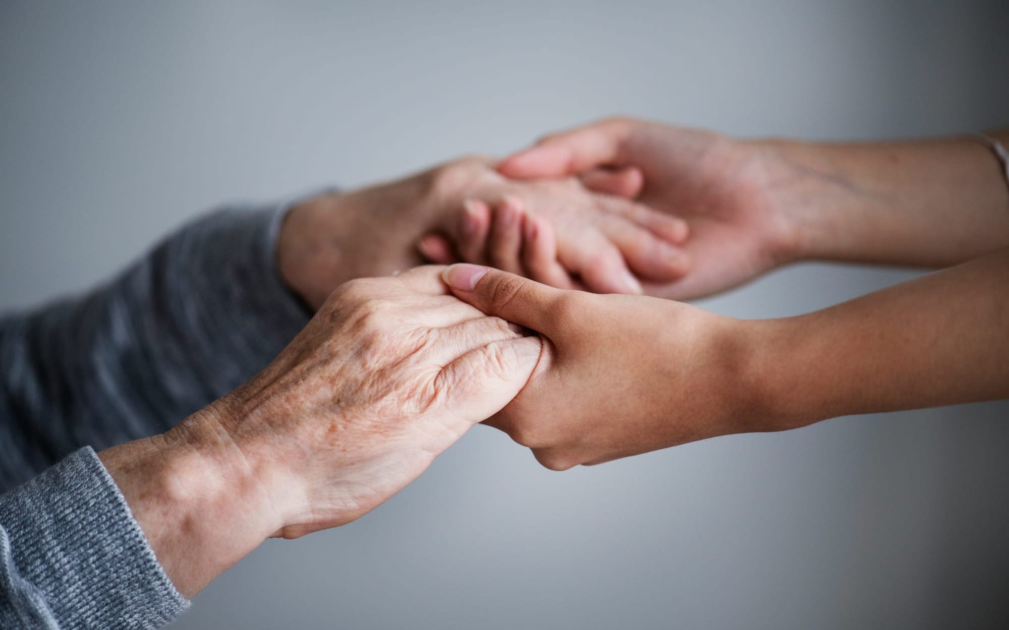 A close-up photograph of a younger person tenderly cradling and holding the weathered, wrinkled hands of an elderly individual against a soft, neutral grey background, conveying deep empathy, comfort, and care.