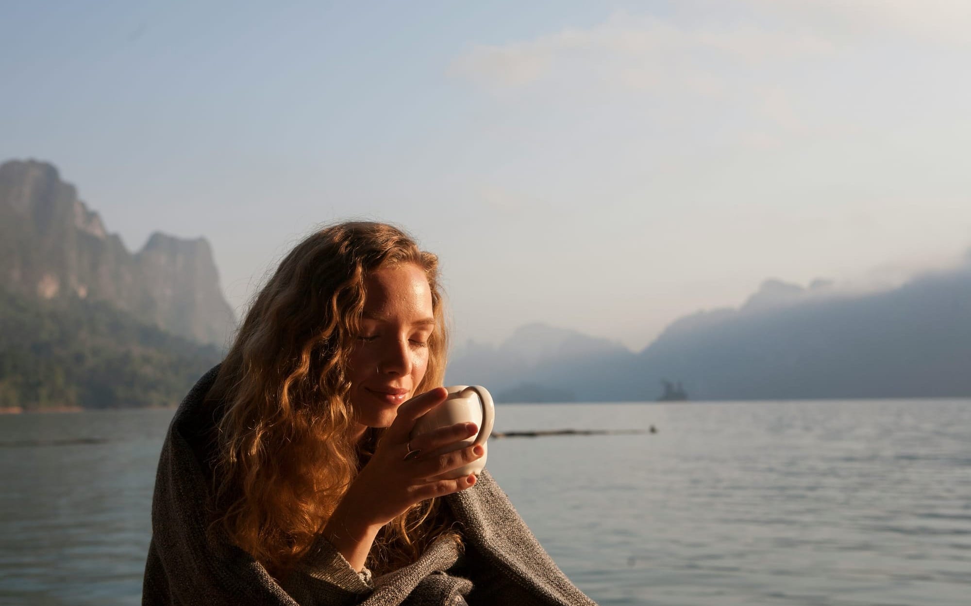 A close-up of a serene woman with her eyes closed holding a warm mug in soft morning sunlight against a calm lake and misty mountains, illustrating daily relaxation rituals.