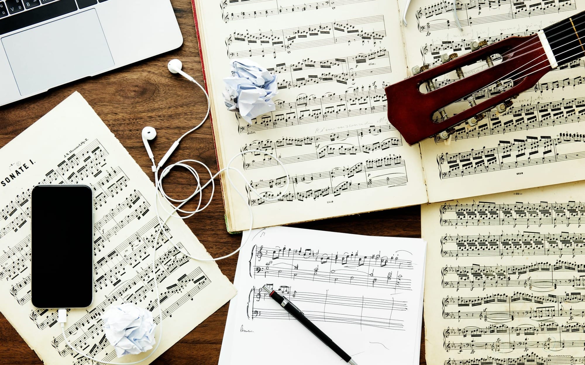 A top-down flat-lay view of a wooden workspace containing a laptop, a smartphone with white wired earbuds, open books of printed sheet music, a guitar neck, handwritten musical notes, and a pen, highlighting the intersection of passive listening and active musical structure.