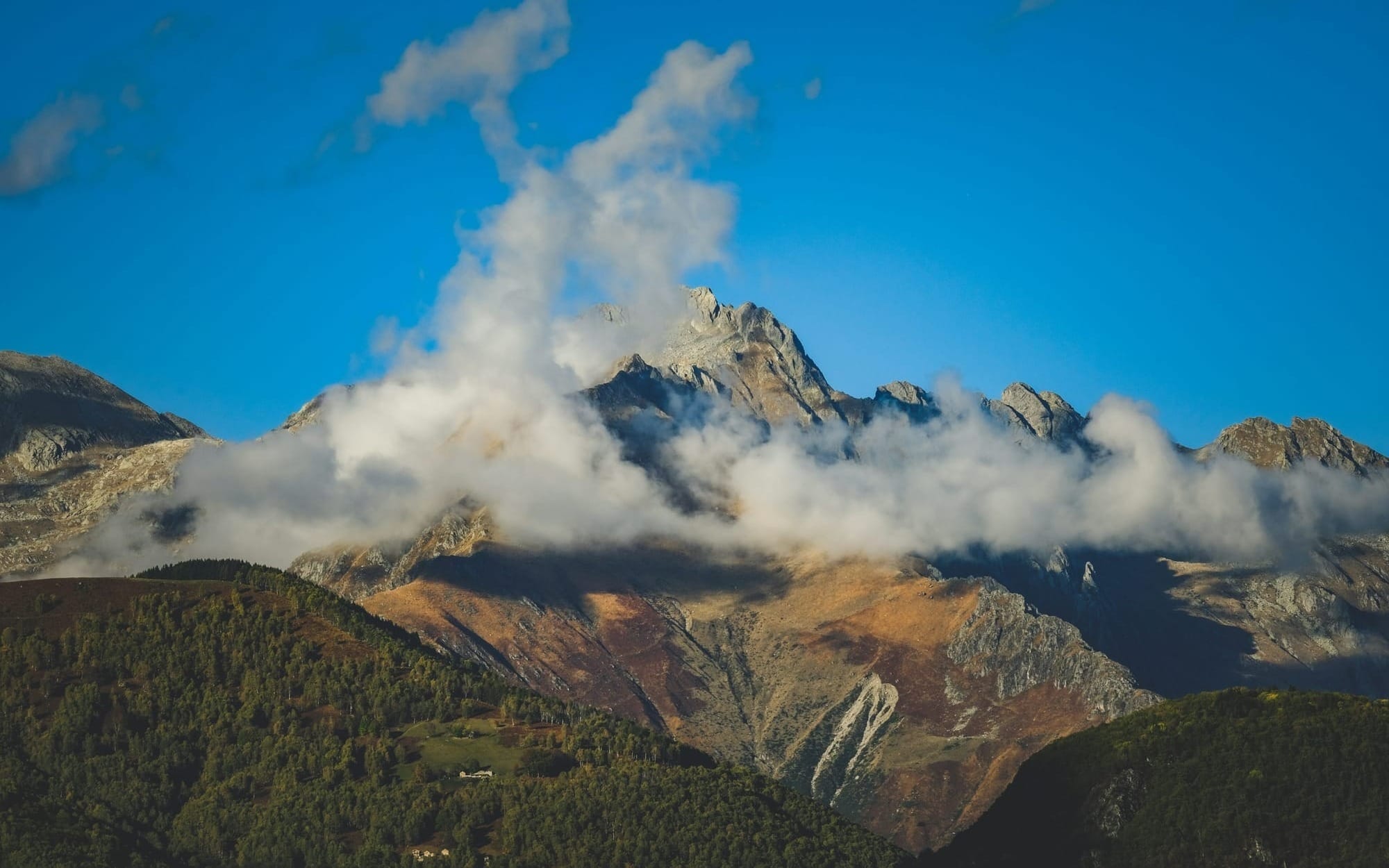 A majestic mountain peak bathed in warm sunlight as low, white clouds begin to clear away under a bright blue sky.