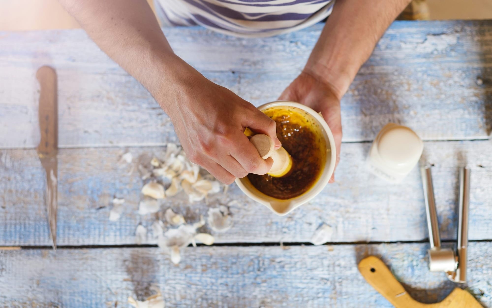 An overhead view of a clinician or herbalist crushing garlic cloves and fresh herbs with a ceramic mortar and pestle on a weathered blue wooden table.