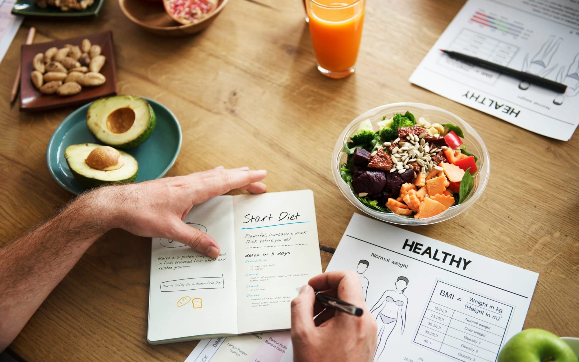An overhead flat lay of a person writing in a food journal on a wooden table surrounded by healthy foods, an avocado, a fresh salad bowl, and functional medicine health tracking sheets.