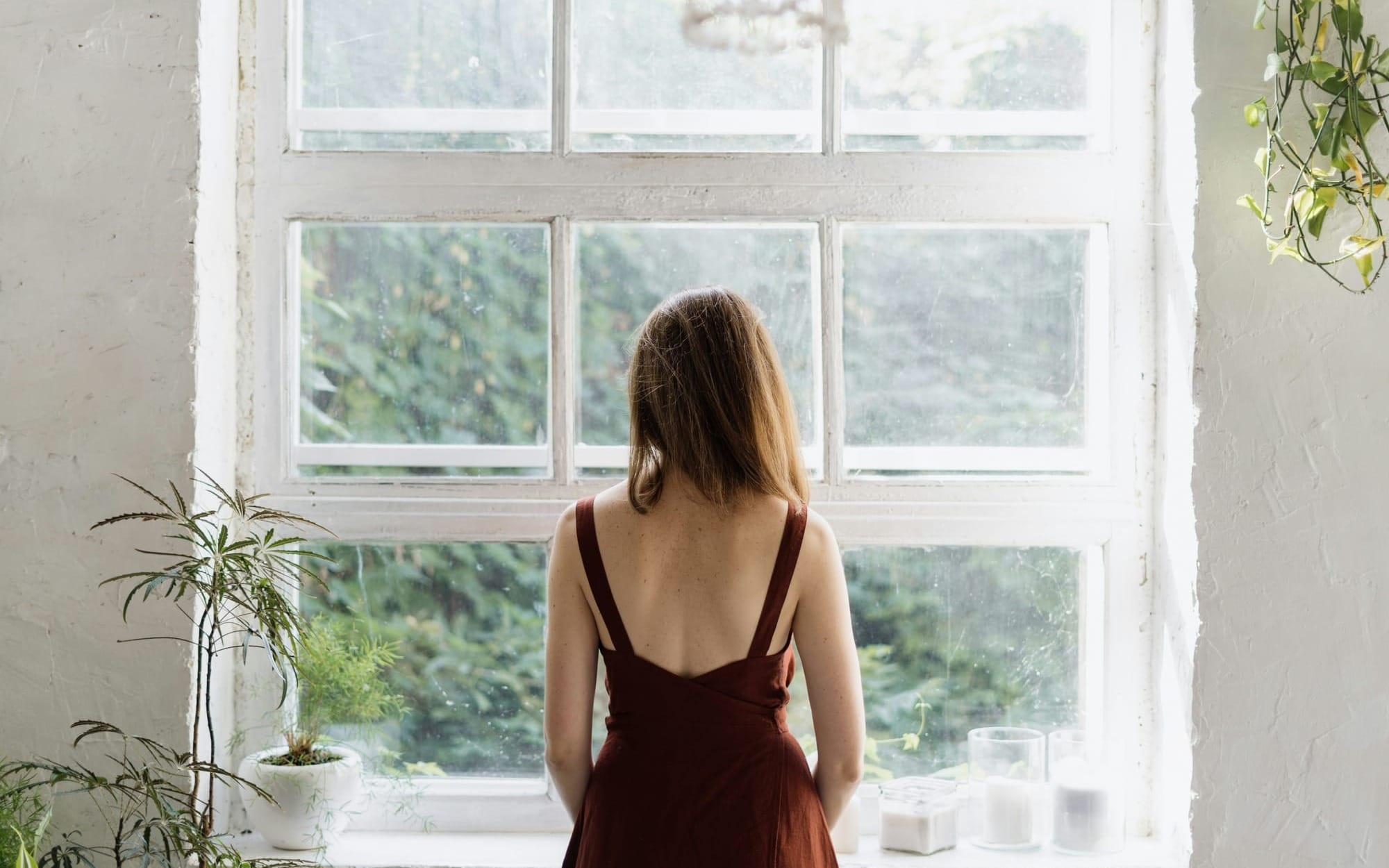 A woman in a rust-colored dress viewed from behind, looking out a large, bright window surrounded by house plants, evoking quiet reflection and mindfulness.