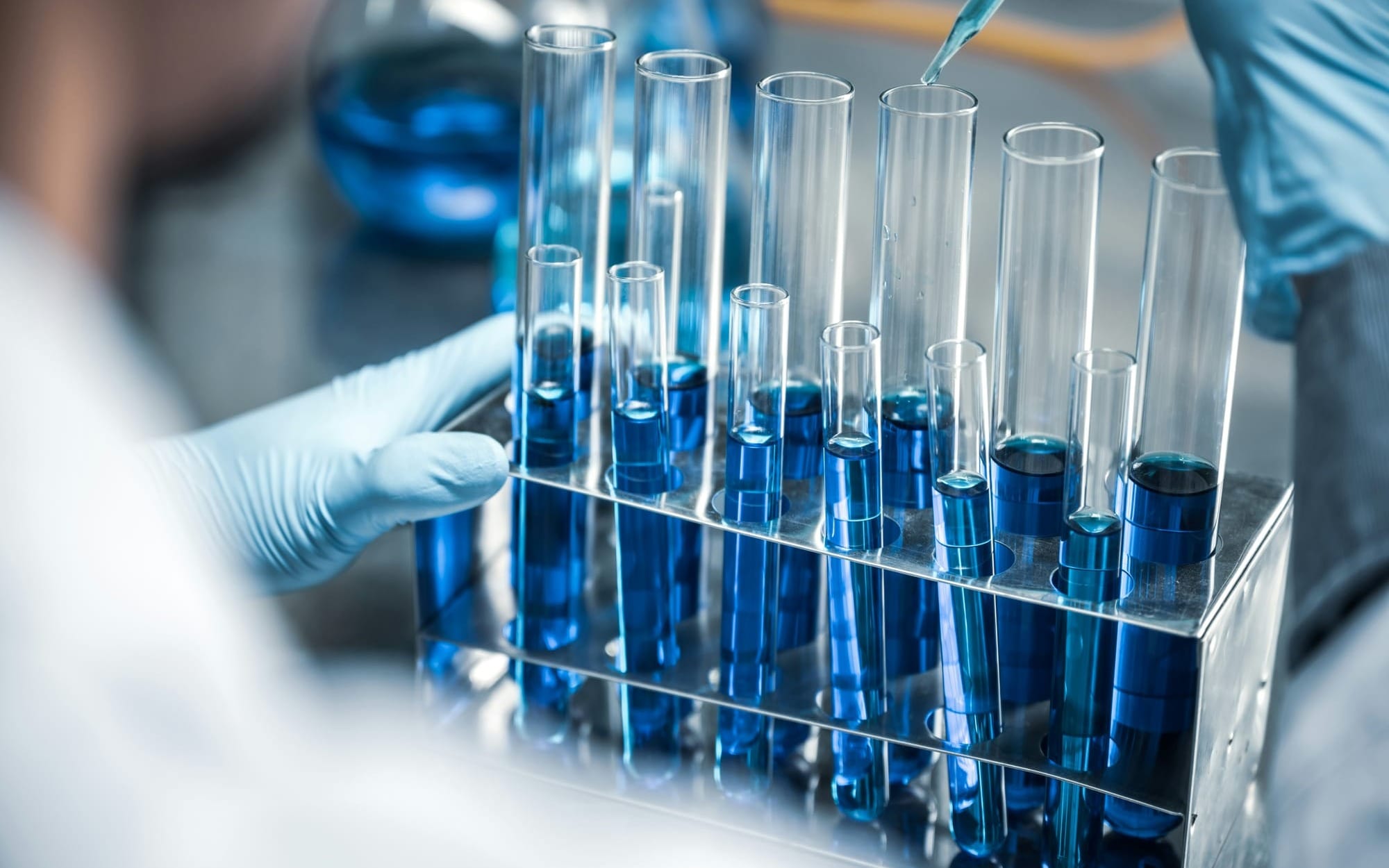 A close-up, selective focus photograph of a metal rack holding multiple glass test tubes filled with dark blue liquid in a clinical laboratory. A gloved hand holds the side of the rack while a pipette drops liquid into a tube, representing conventional medical diagnostic testing.