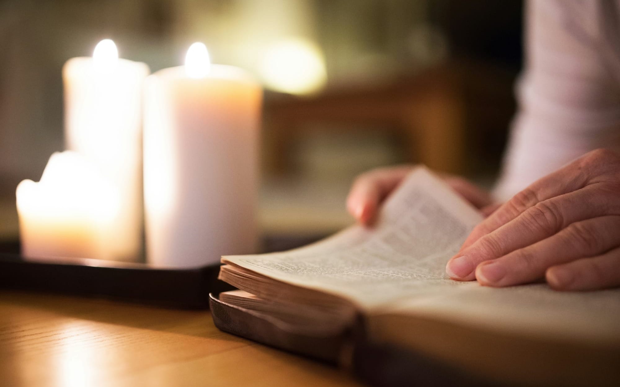 A close-up shot of a person’s hands gently turning the pages of an open book on a wooden table, illuminated by the warm, soft glow of thick white candles in the background.