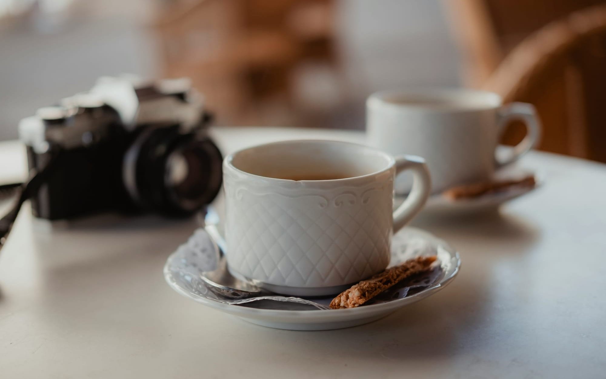 An intimate, shallow depth-of-field photograph of two white ceramic coffee cups on a table with a vintage film camera resting blurred in the background, metaphorically representing shared family moments and the preservation of fading memories.