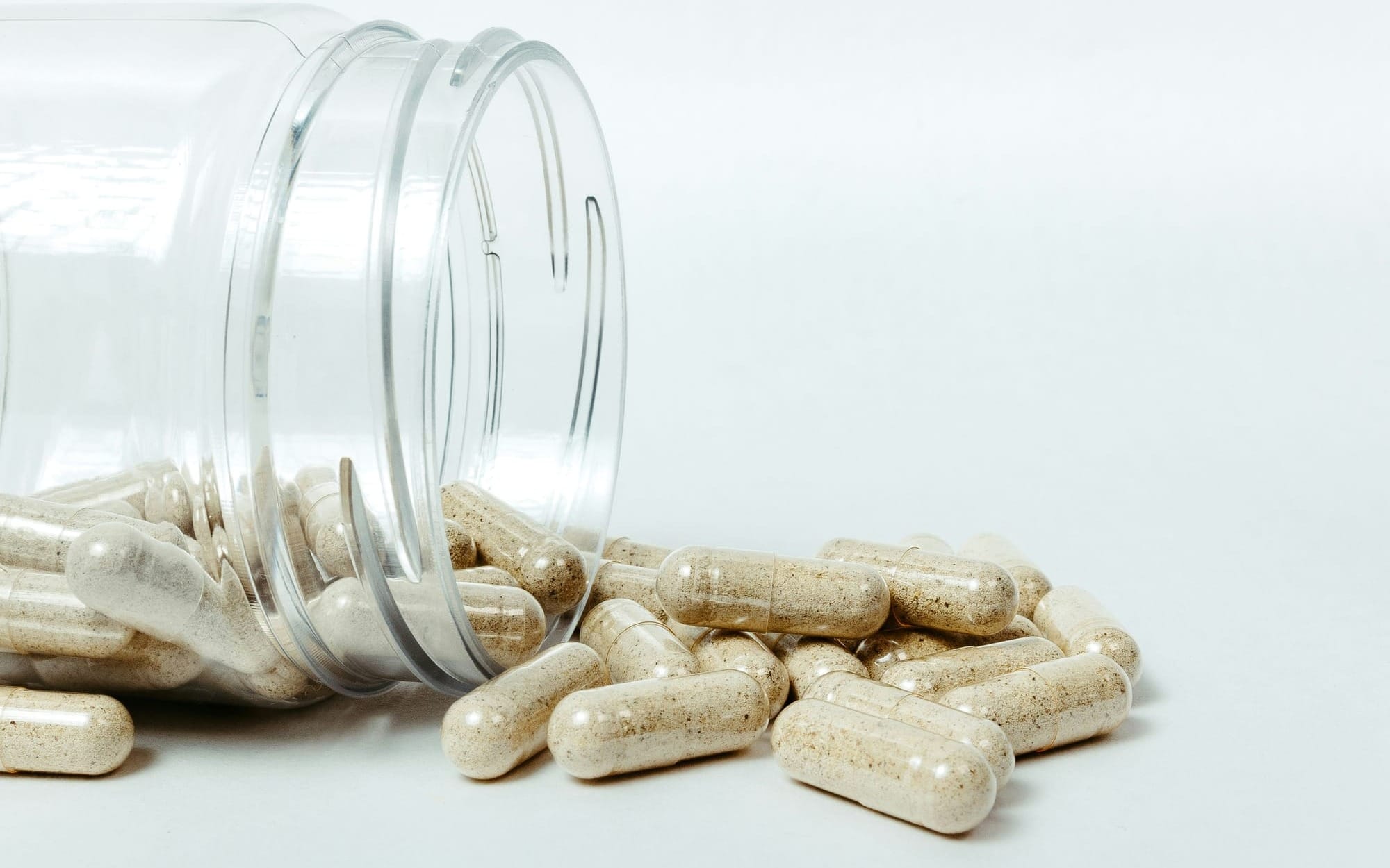 A clean, high-angle studio photograph of transparent supplement capsules filled with natural herbal powder spilling from a clear bottle onto a white surface, representing a structured Functional Medicine treatment protocol.