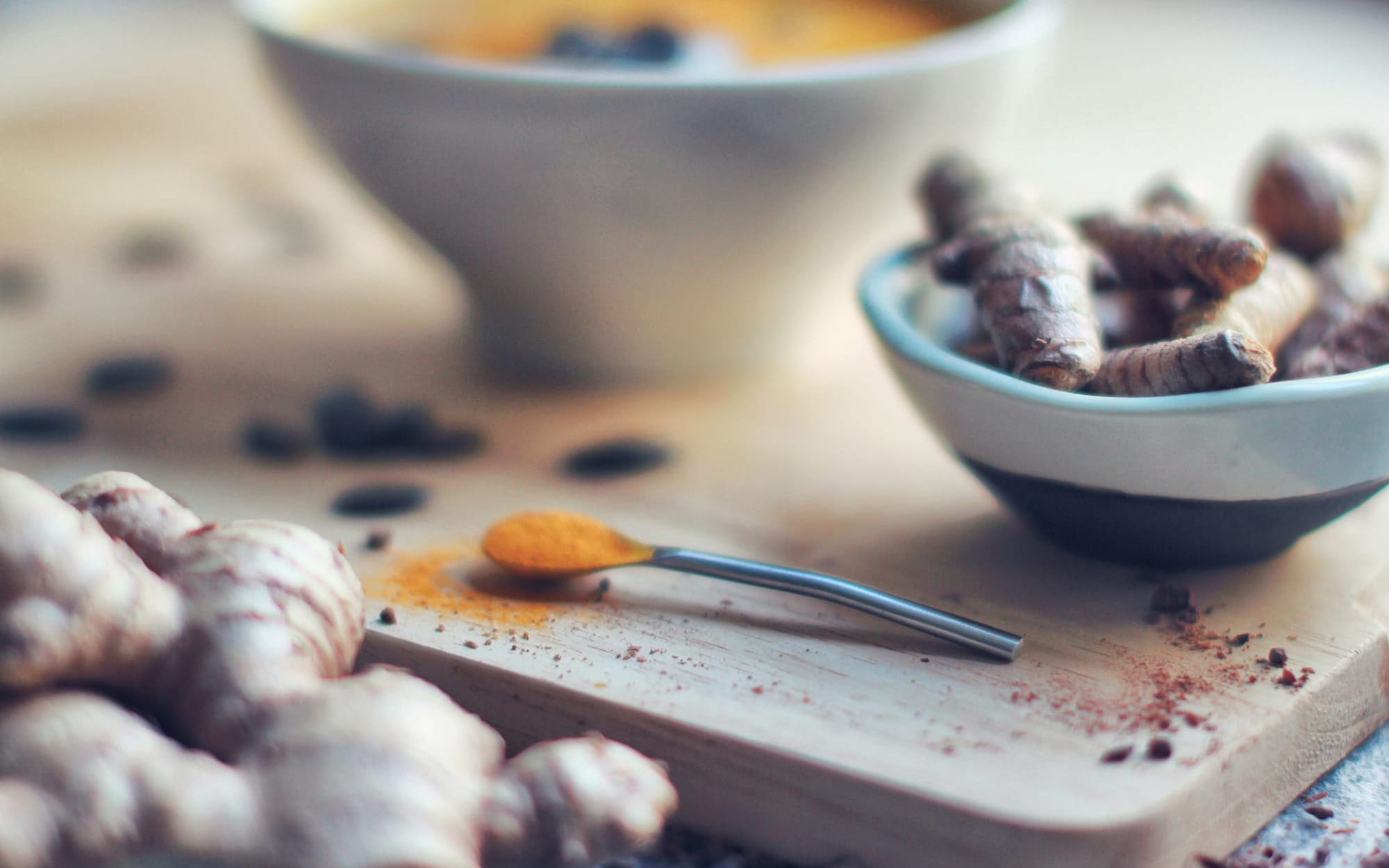A close-up culinary scene featuring fresh ginger rhizomes, a small bowl of raw turmeric roots, and a silver spoon filled with golden turmeric powder on a wooden cutting board.