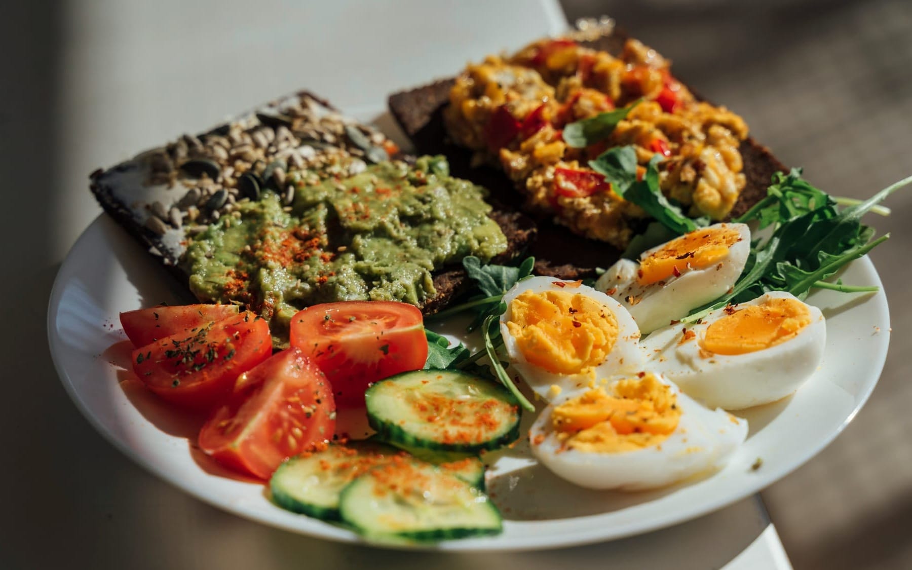 A savory breakfast plate featuring sliced hard-boiled eggs, scrambled eggs on dark rye bread, avocado, and fresh tomatoes, representing nutrient-dense, choline-rich foods.