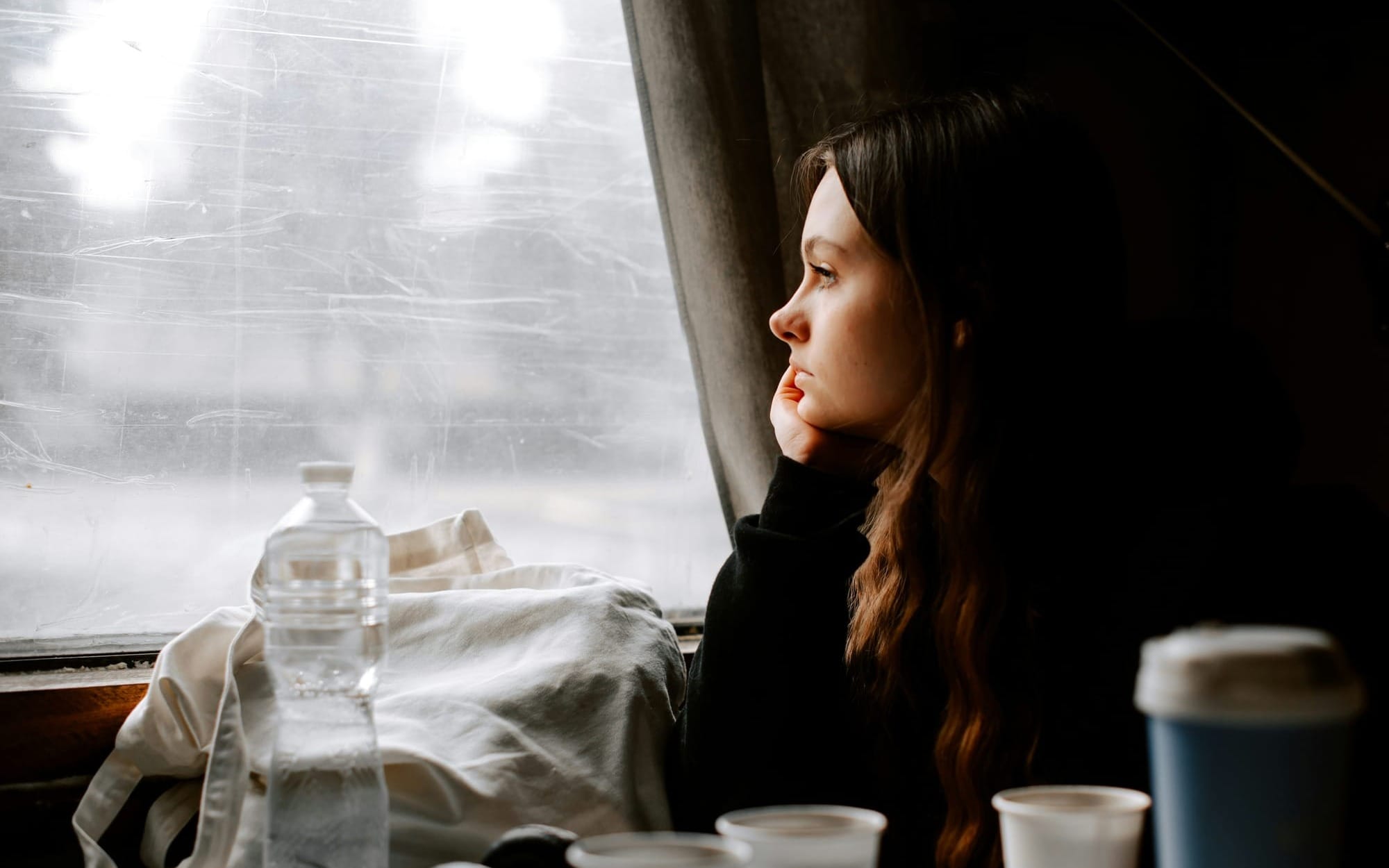 A contemplative young woman looking thoughtfully out a window in dim, cinematic lighting, reflecting clinical frustration and exhaustion.