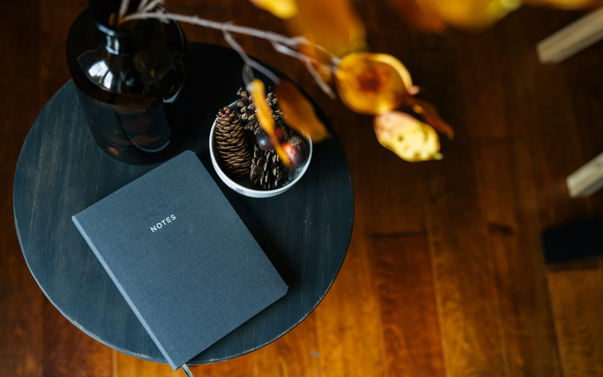 A premium, top-down view of a dark grey notebook titled Notes on a black round side table, surrounded by warm autumn foliage and wooden elements, symbolizing a grounding wellness ritual.
