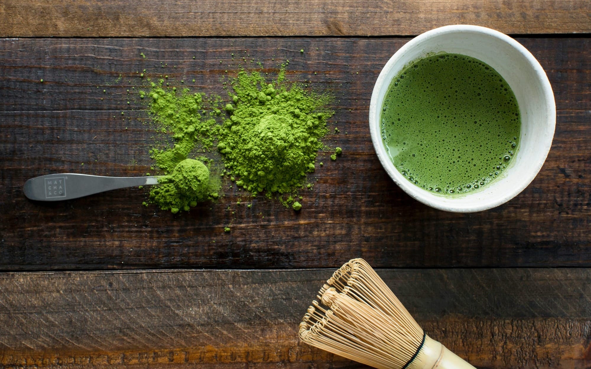 An overhead flat lay of green matcha tea powder, a traditional bamboo whisk, and a ceramic cup of whisked green tea on a dark wooden background.