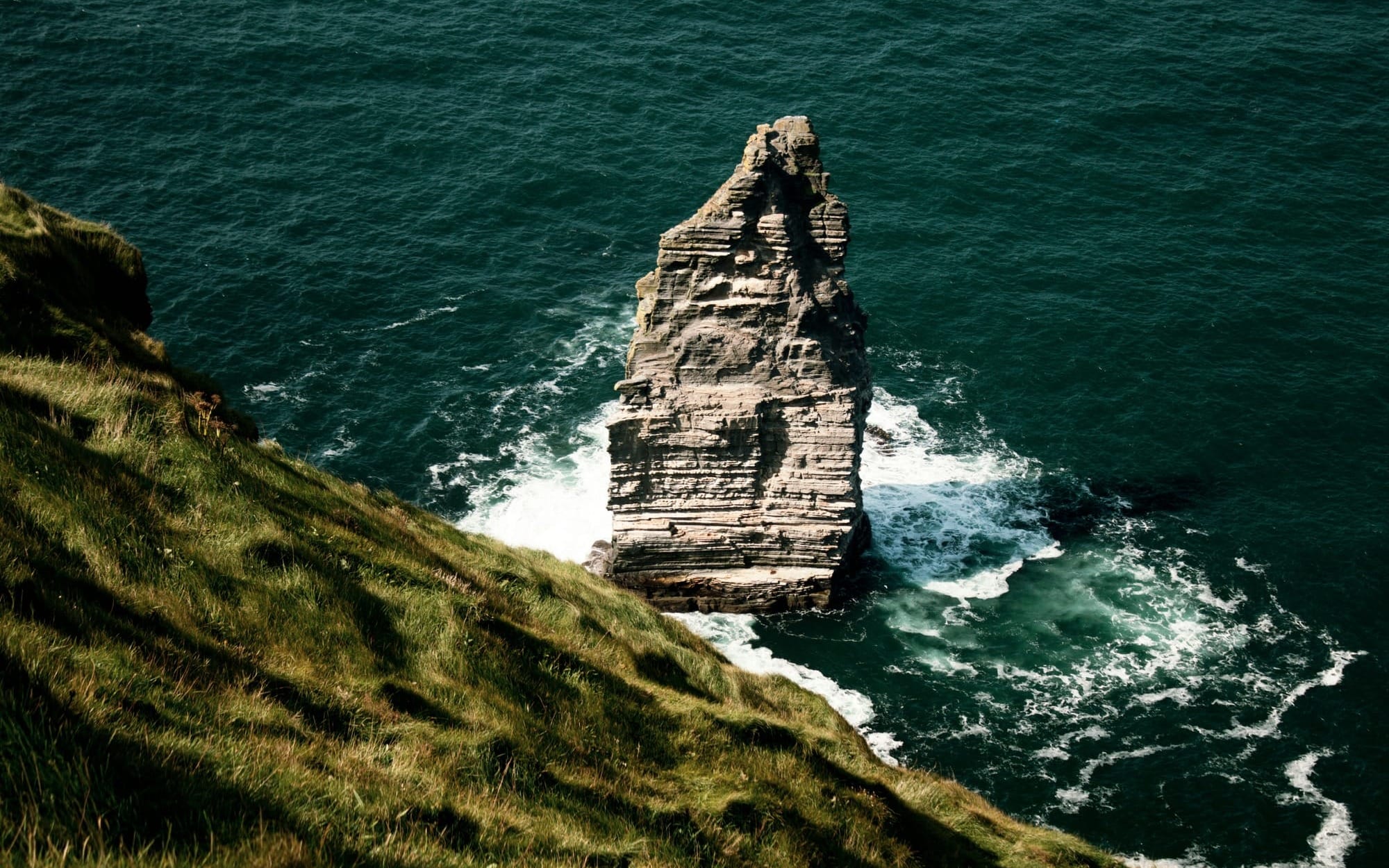 A striking high-angle view of a massive rock sea stack standing firm in rough teal ocean waters next to a green grassy cliff side, symbolizing deep internal strength and stability.
