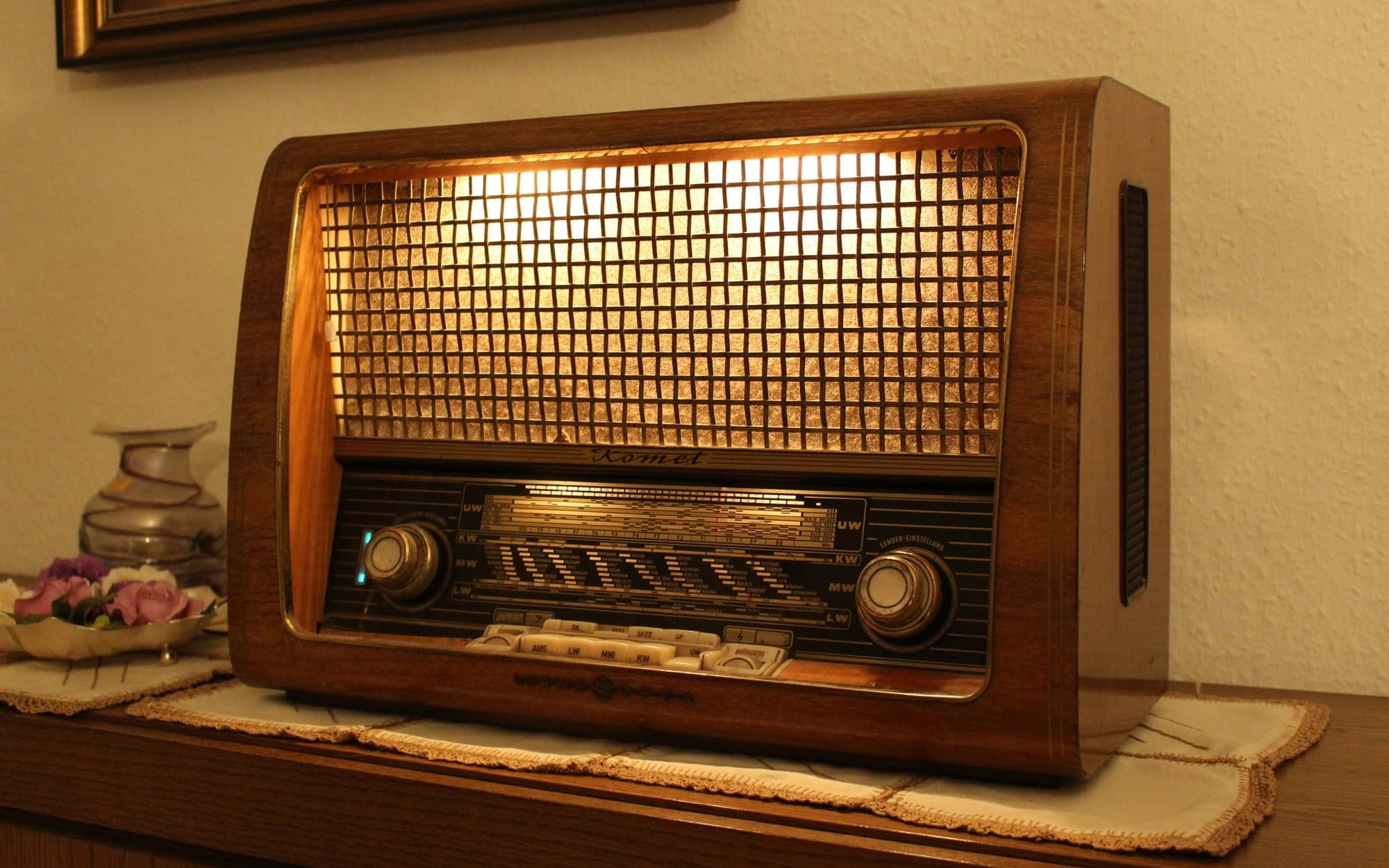 A vintage wooden Komet tube radio sitting on a table with its golden dial and mesh grille warmly illuminated from within, serving as a visual metaphor for long-held musical memories in cognitive care.