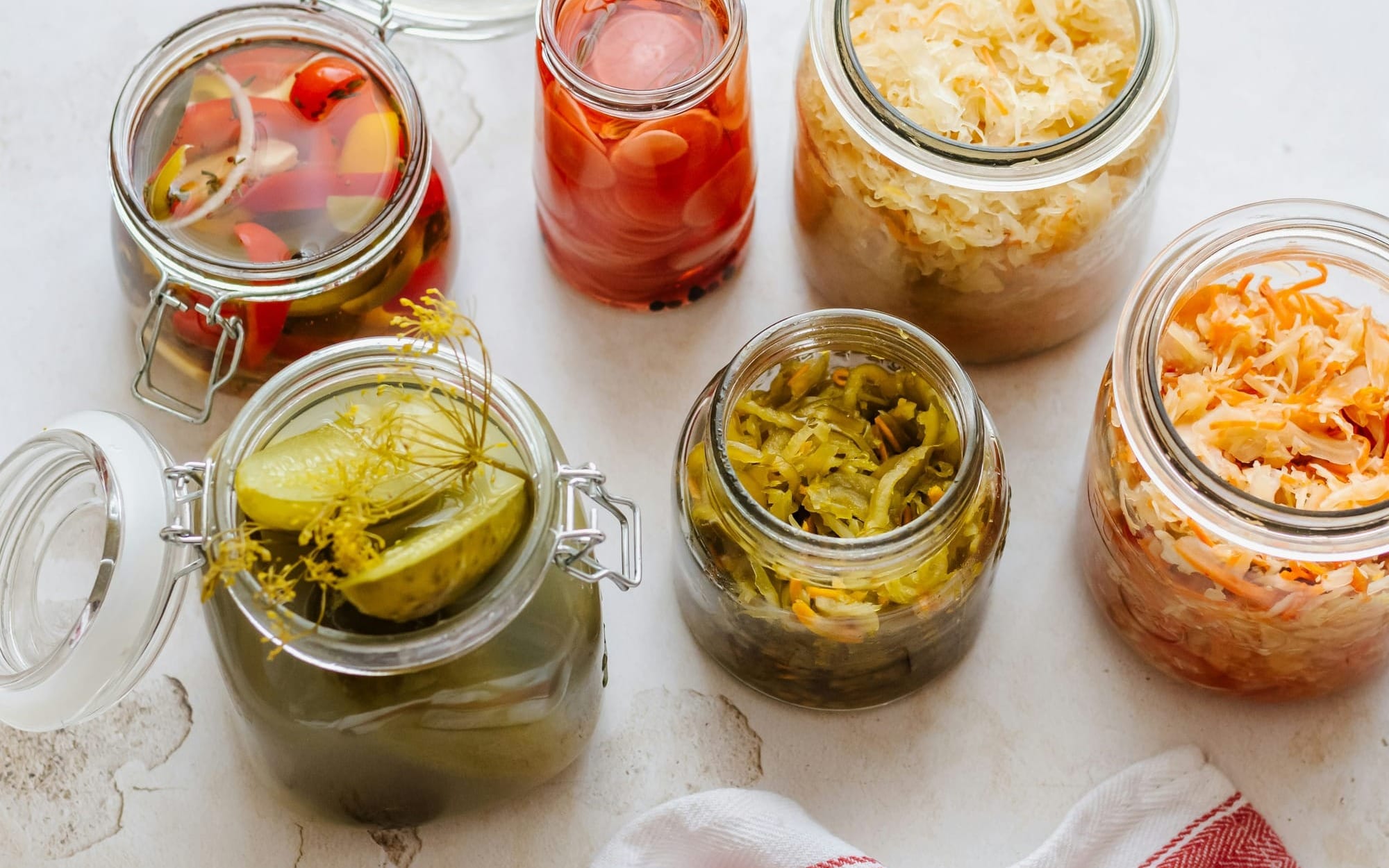 An overhead view of open glass jars filled with fermented probiotic foods including sauerkraut, pickled cucumbers, radishes, and carrots on a bright surface.