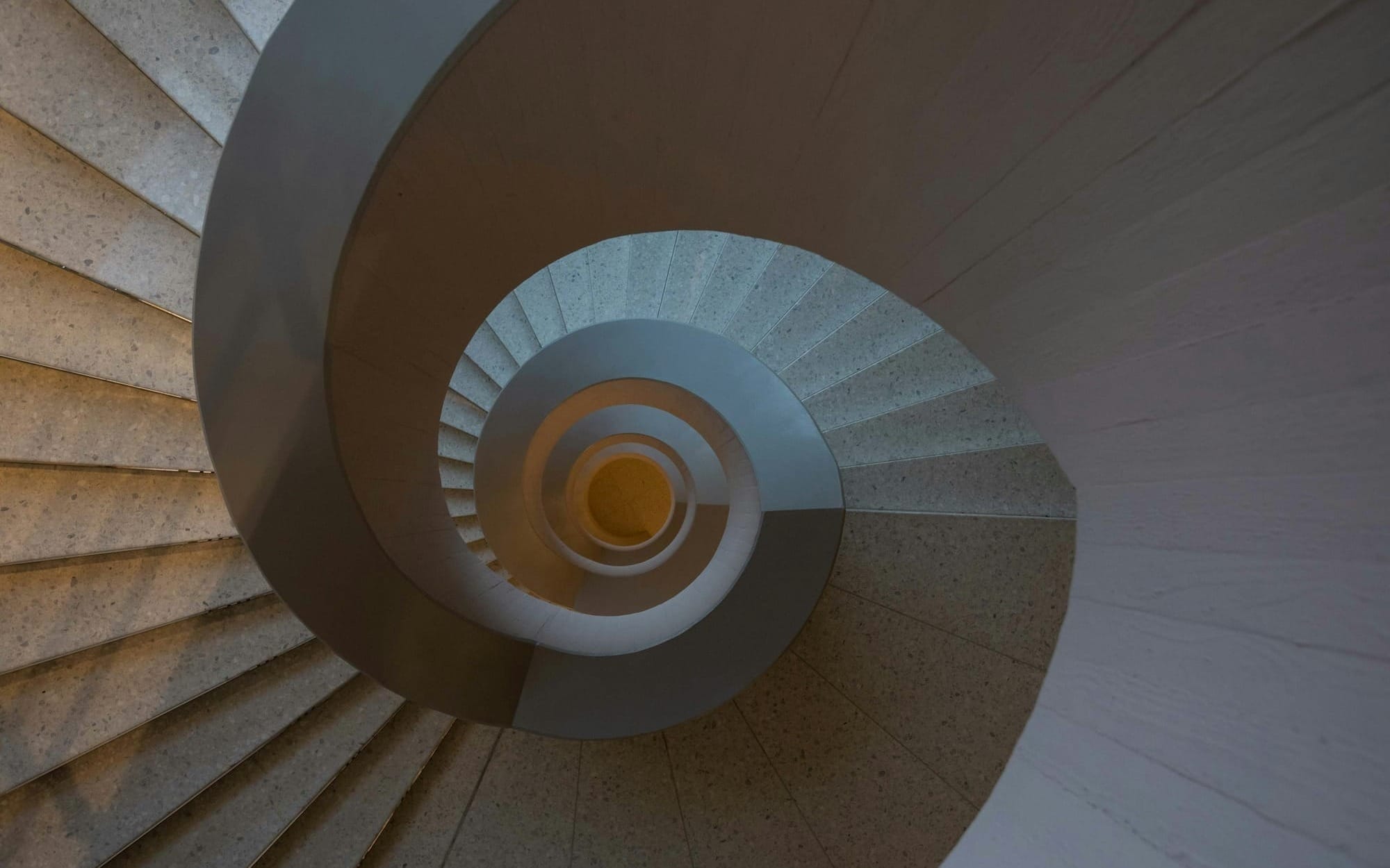 An abstract, top-down geometric view looking into a winding concrete spiral staircase that curves downward into a deep center.