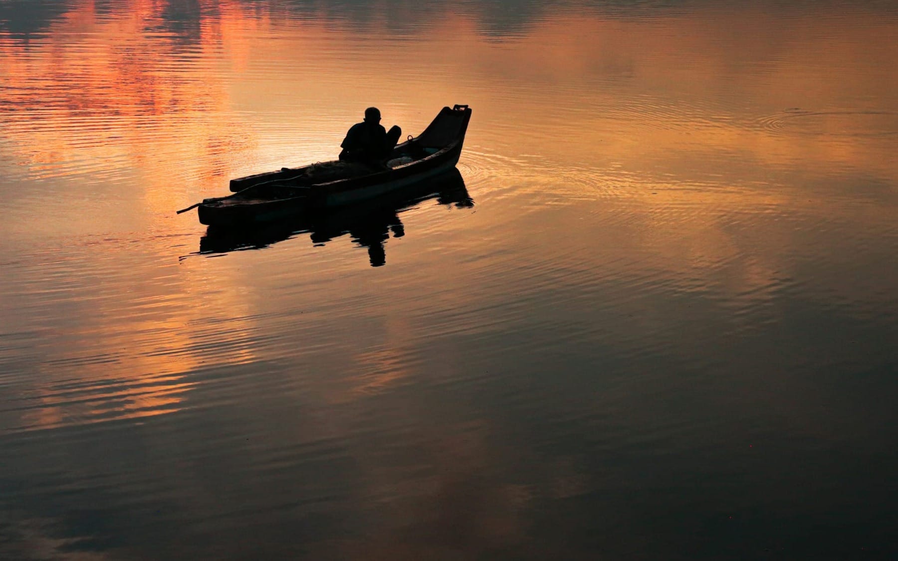 A silhouette of a lone fisherman sitting in a small wooden boat on calm, glassy water, reflecting a vibrant orange and pink sunset sky.