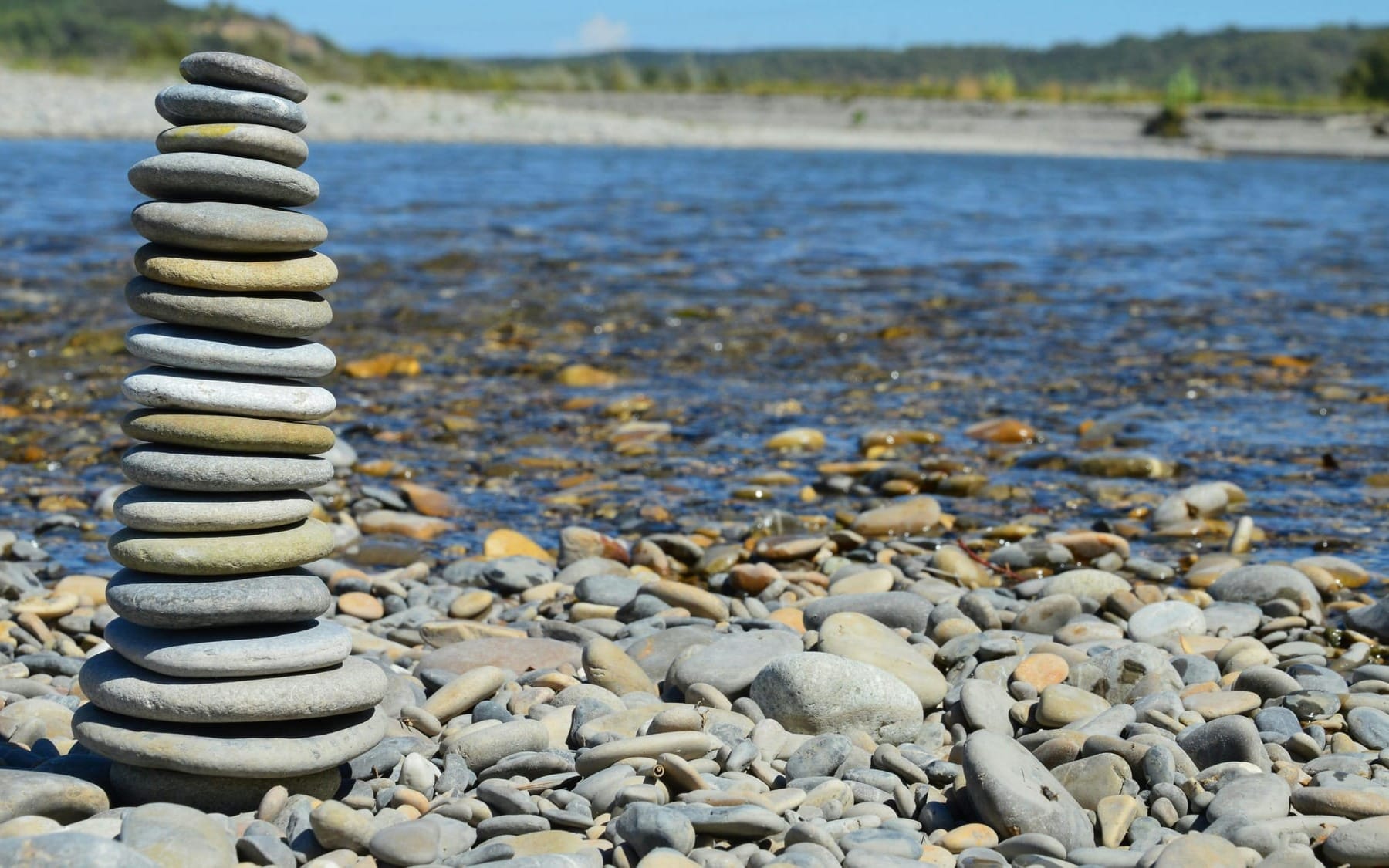 A tall stack of smooth, grey river stones perfectly balanced in a tower on a rocky shoreline next to a wide, flowing river under bright daylight.