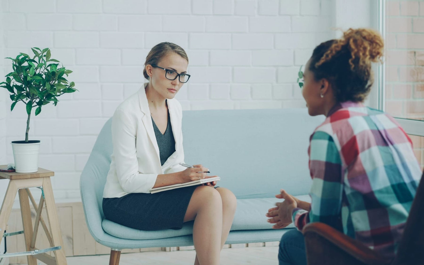 An integrative health coach in a white blazer actively listening and taking notes during a collaborative wellness consultation with a client in a bright, modern office.