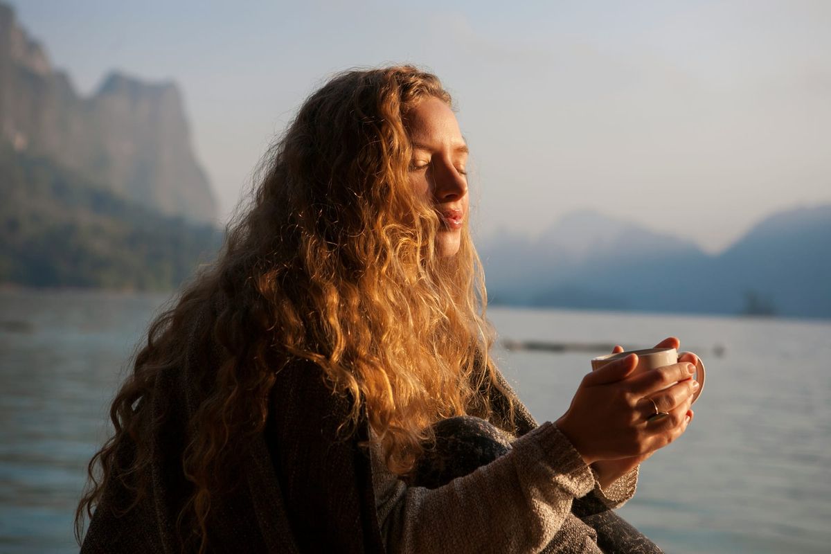 A serene, close-up photograph of a woman with long, curly hair sitting by a calm lake at sunrise. Her eyes are closed, her face is turned toward the warm golden sunlight, and she is holding a