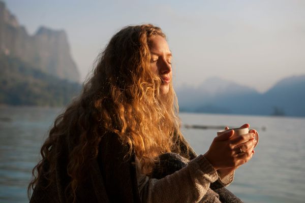 A serene, close-up photograph of a woman with long, curly hair sitting by a calm lake at sunrise. Her eyes are closed, her face is turned toward the warm golden sunlight, and she is holding a