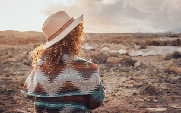 A woman wearing a wide-brimmed hat and a patterned poncho stands with her back to the camera, looking out over a vast, open landscape at sunset, evoking a sense of self-acceptance.