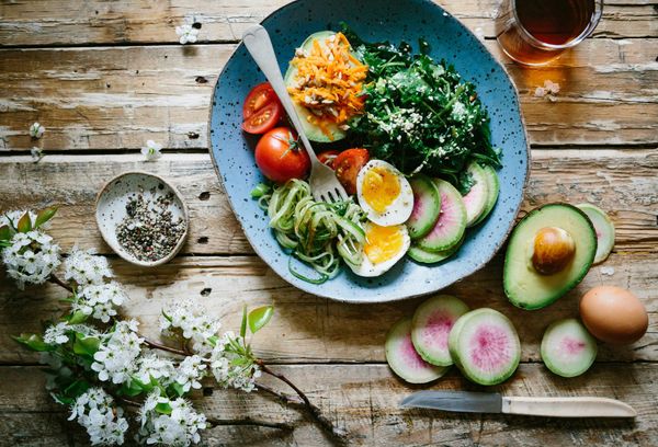 A healthy, nutrient-dense meal featuring a colorful salad bowl with sliced avocado, boiled eggs, tomatoes, shredded carrots, and leafy greens on a rustic wooden table with white blossoms.