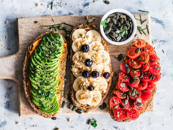 Three slices of artisan whole-grain toast topped with avocado, bananas, and cherry tomatoes on a wooden cutting board, representing functional nutrition and a healthy vagus nerve diet.