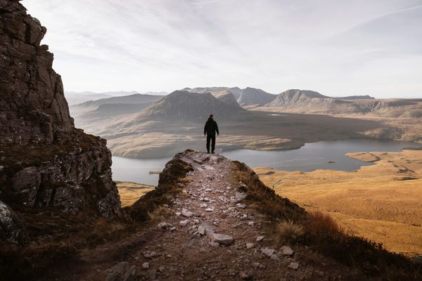 A lone silhouette of a person standing on a rocky mountain ridge, looking out over a vast landscape of misty peaks, hills, and a calm lake under a soft sky.