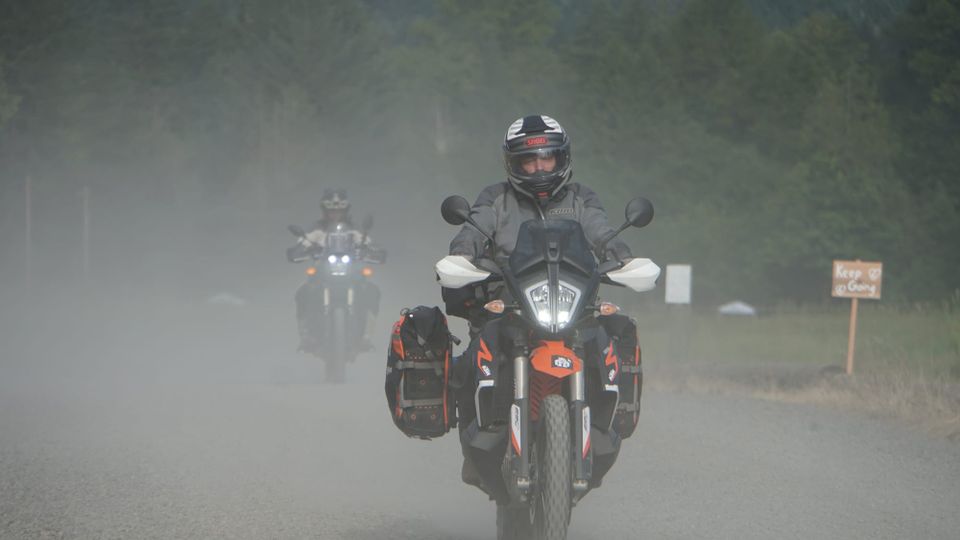 An adventure motorcycle with a handsome rider travels down a dusty road. A second ride follows behind.