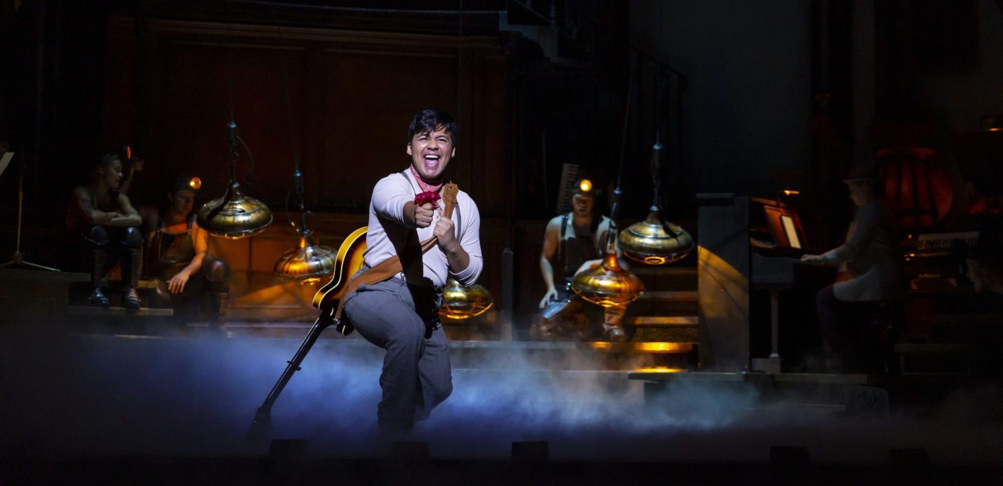 A man clutching a red rose, guitar slung across his back, sings while surrounded by eerie smoke and miners wearing headlamps.