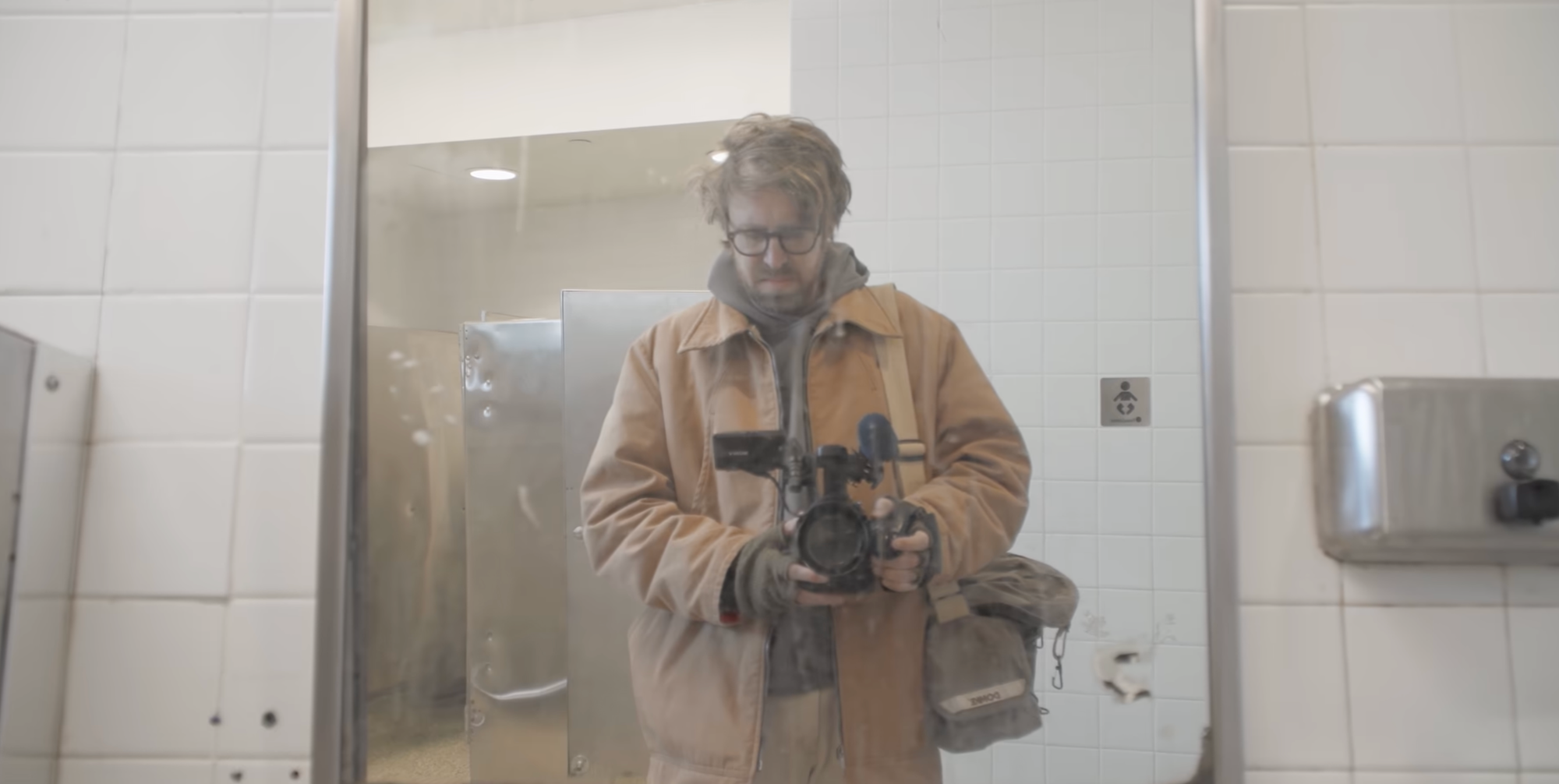 John Wilson holding his handheld camera in a dirty bathroom mirror