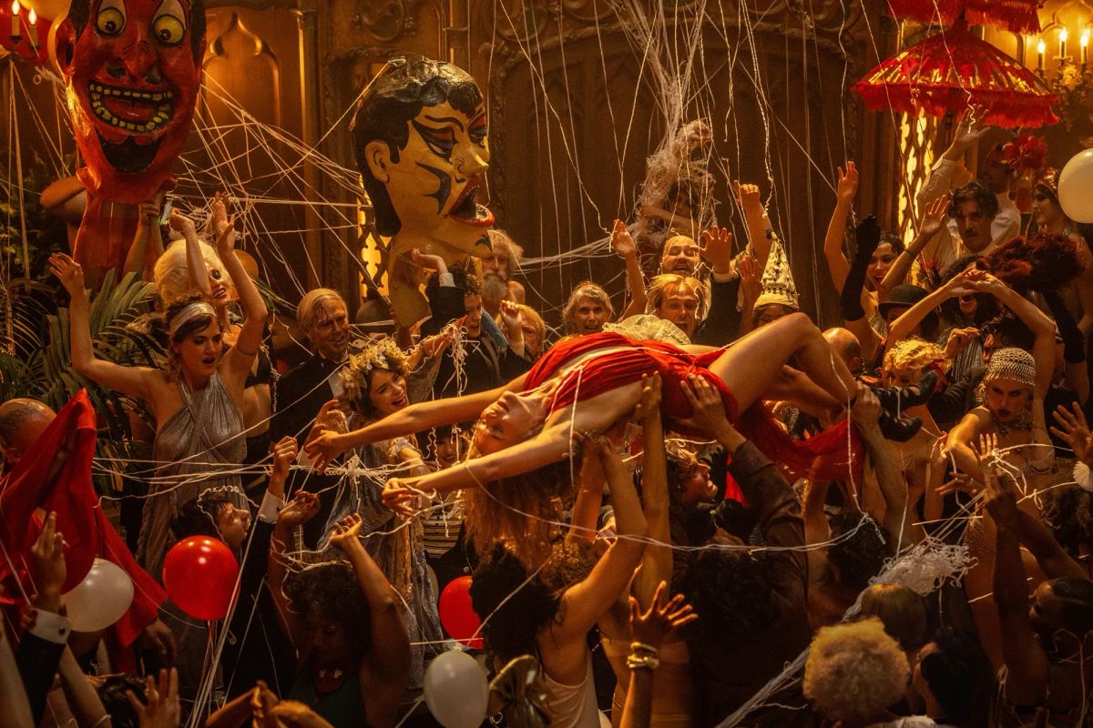 Margot Robbie, in a red dress, rides atop a throng of people, borne up by their hands, at a massive party.