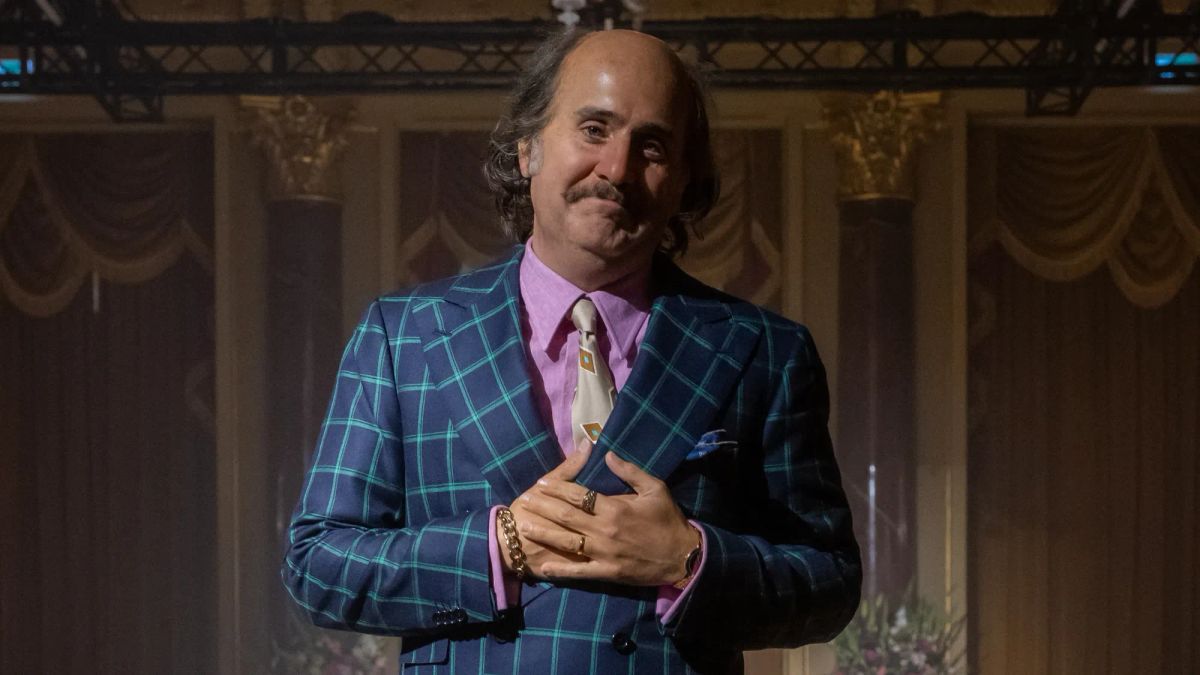 Jared Leto has a lot of padding added to his face via makeup in House of Guc. He wears a bald cap and a blue and green suit.