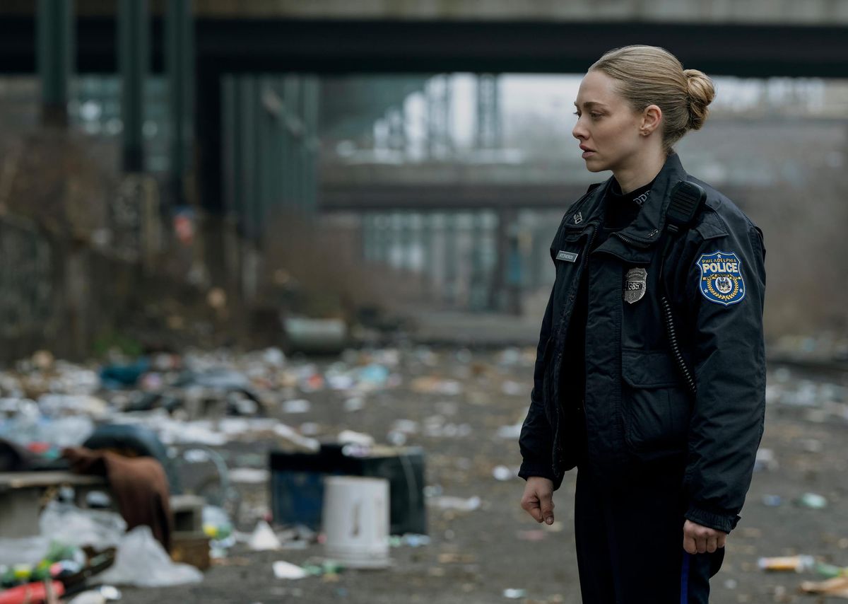 Amanda Seyfried in a Philadelphia police uniform stares with concern at a messy area covered in trash.