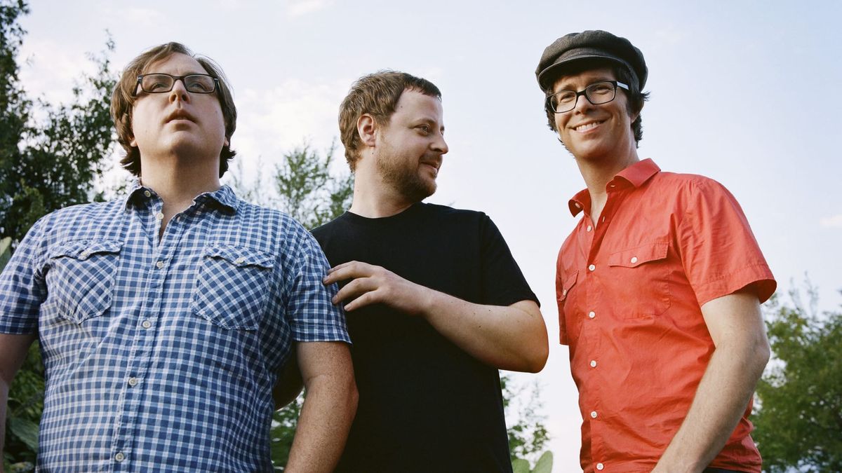 The members of Ben Folds Five pose in front of a forested area. Ben Folds looks at the camera; the other two look away.