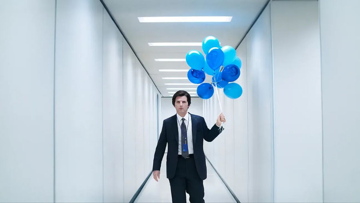 Mark S. (Adam Scott) is in a suit. He stands in a long, featureless, white hallway. He holds blue balloons with his face on them.