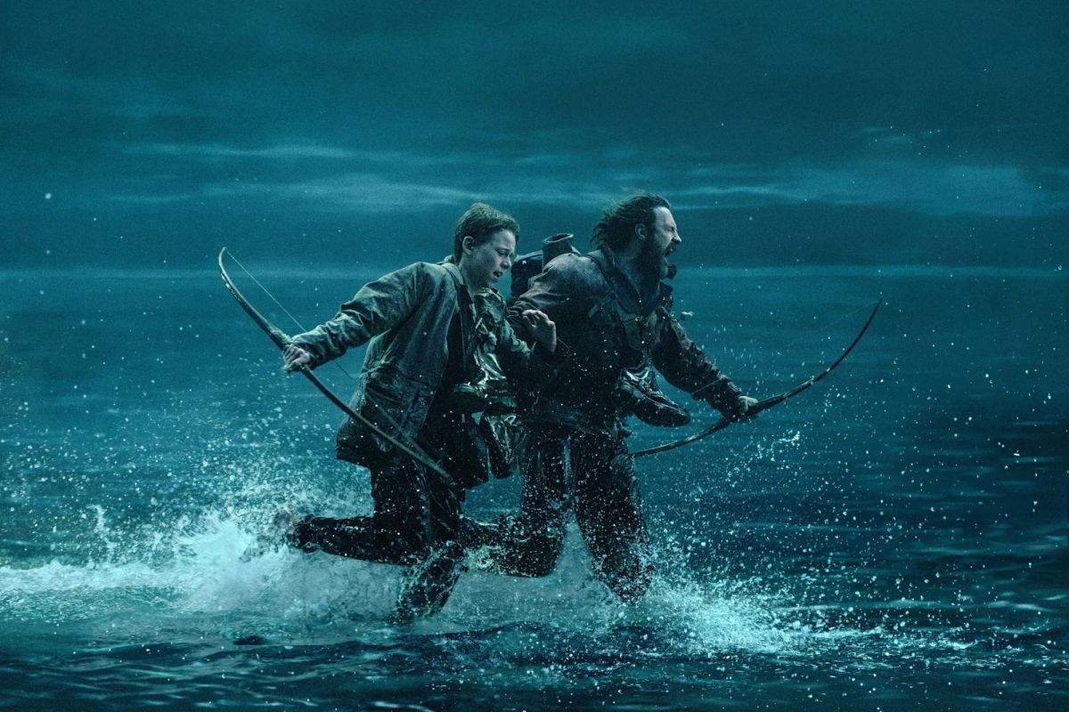 A boy and a man, both holding bows, crash through shin-deep water, running away from something. It's nighttime.