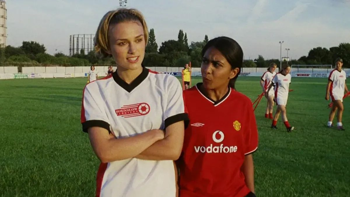 Two young women walk across a soccer pitch in their jerseys. One is tall and white, the other shorter and of Indian descent.