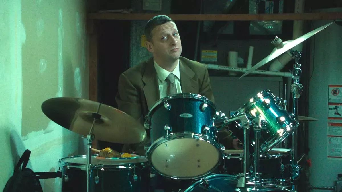 Tim Robinson, clad in a deeply tan suit, sits behind a sweet drum set.